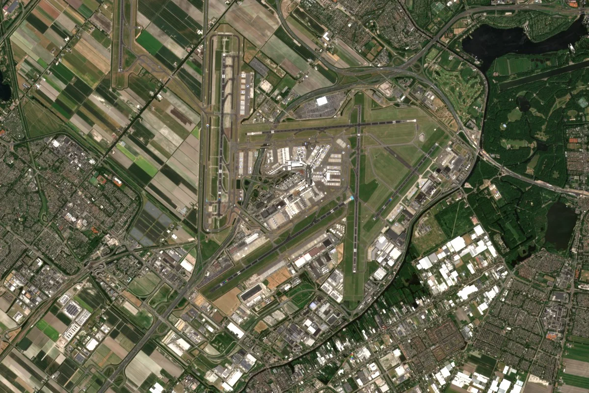 A large airport among green fields and small towns, with several long runways, taxiways, terminal buildings and aprons where parked aircraft show only as small pale shapes.