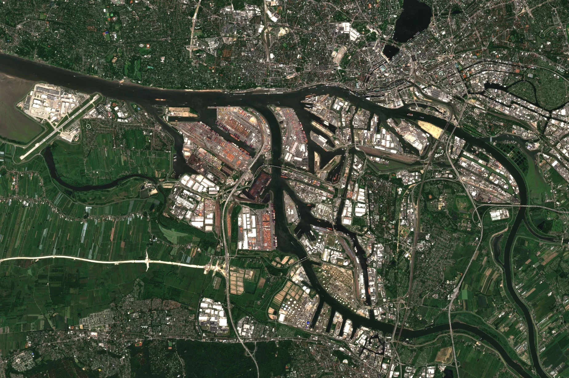 A large river port seen from above. A wide river crosses the image and splits into dark harbour basins between long quays, some lined with coloured containers, with clusters of round white storage tanks, ships moored alongside, farmland to the south and west and dense city streets to the north-east.