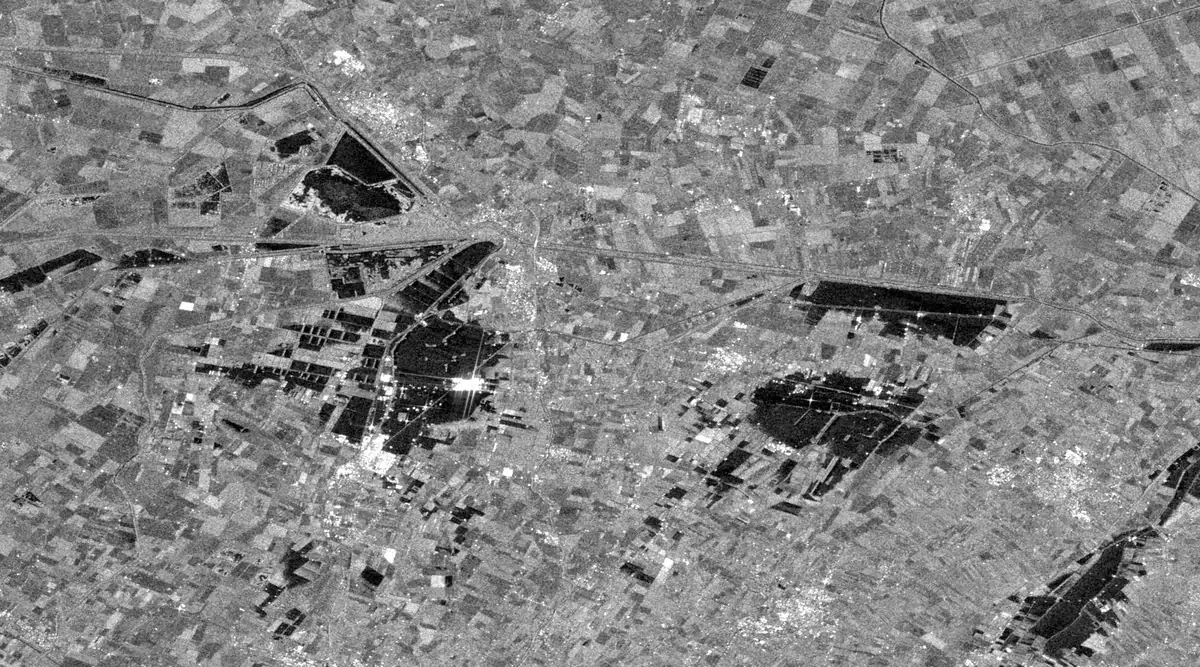 A grey radar image of flat farmland made of many small rectangular fields, with several large black areas of flood water spreading across the fields around a small town in the left centre and in the middle right.