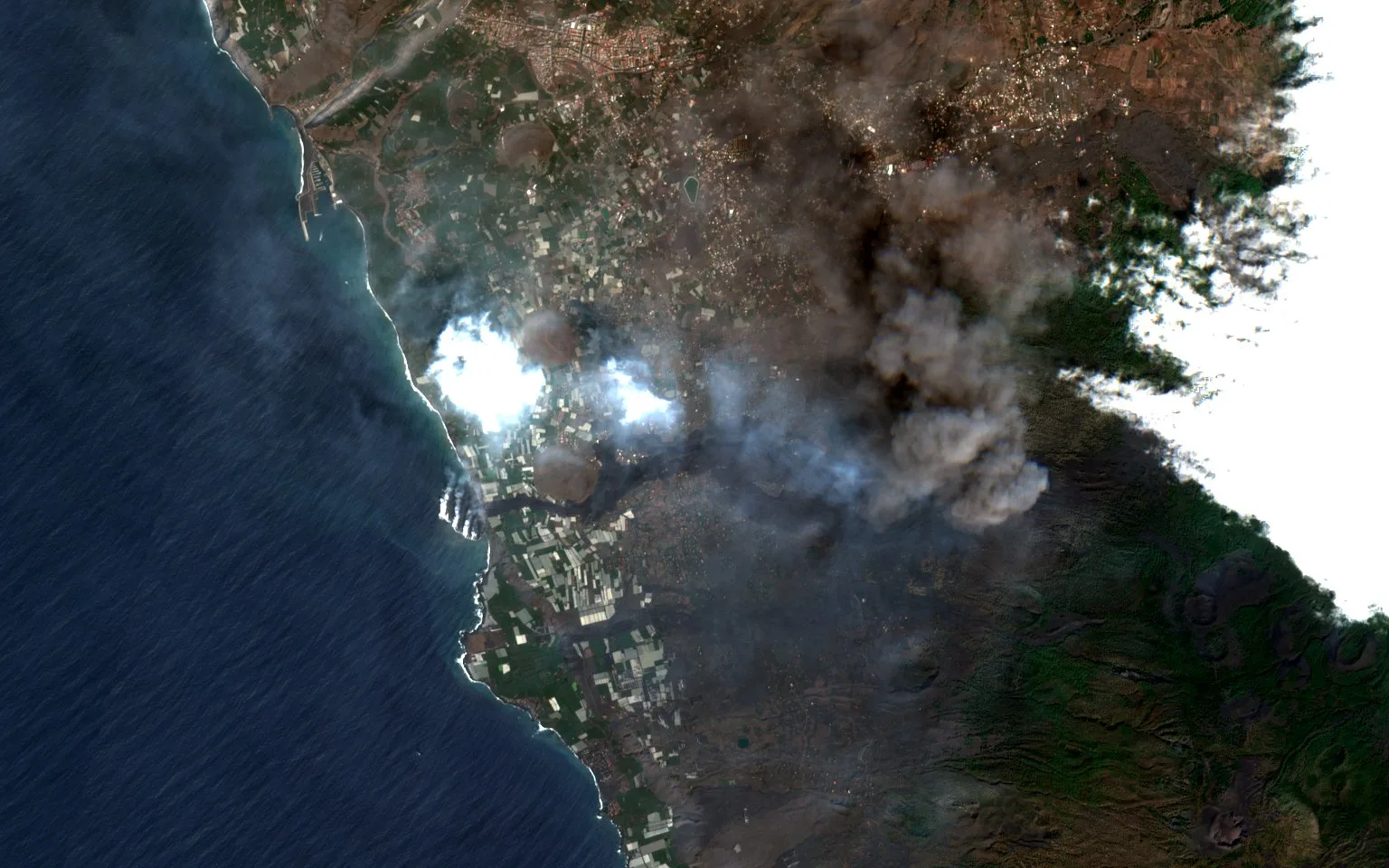 The same coast eleven days into the eruption, with a dark ash plume over the hills, white cloud, and the first lava flow running down to the sea