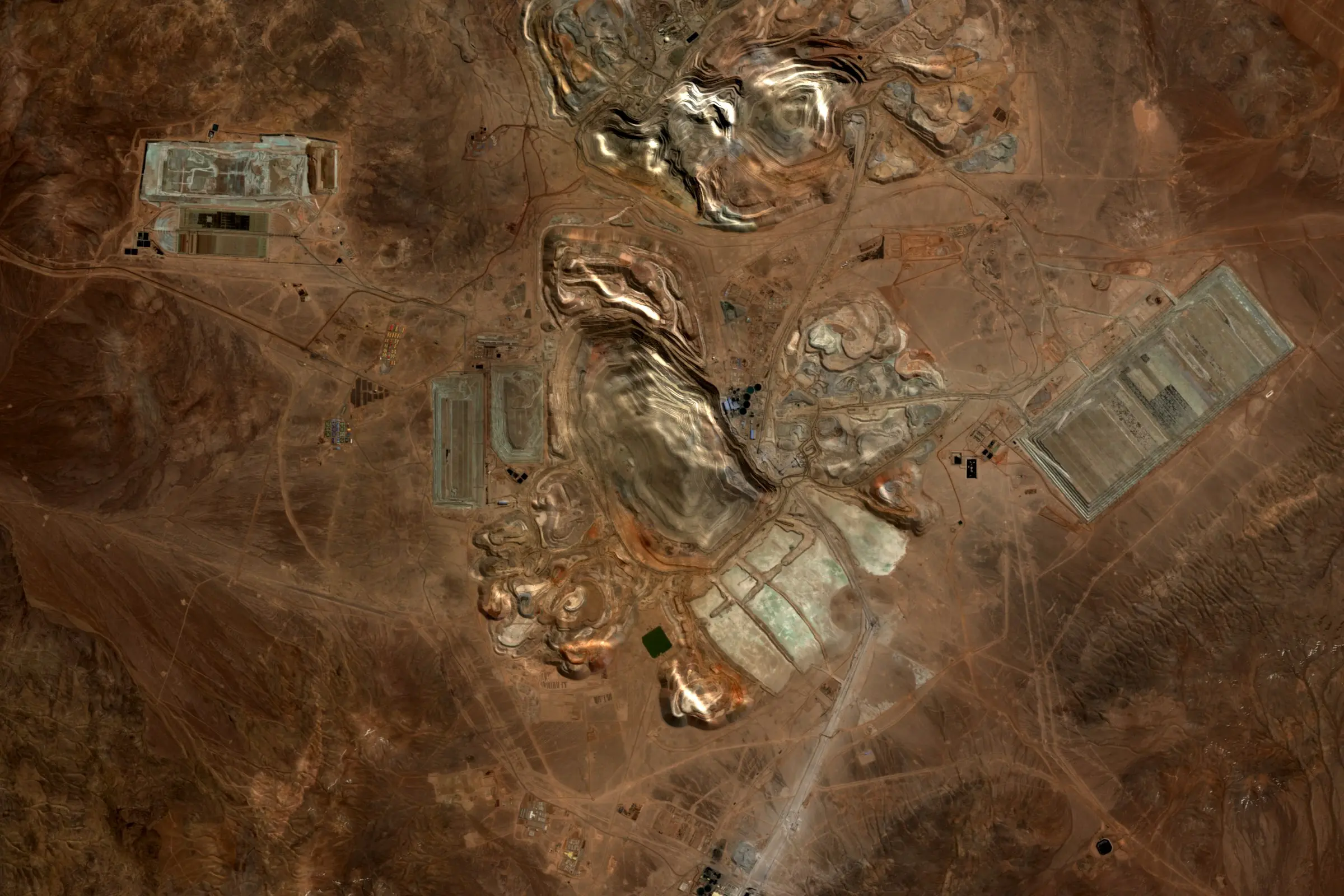 Satellite view of the Escondida copper mine in the Atacama desert, Chile, with pale open pits, waste dumps and processing plant on rust-brown desert