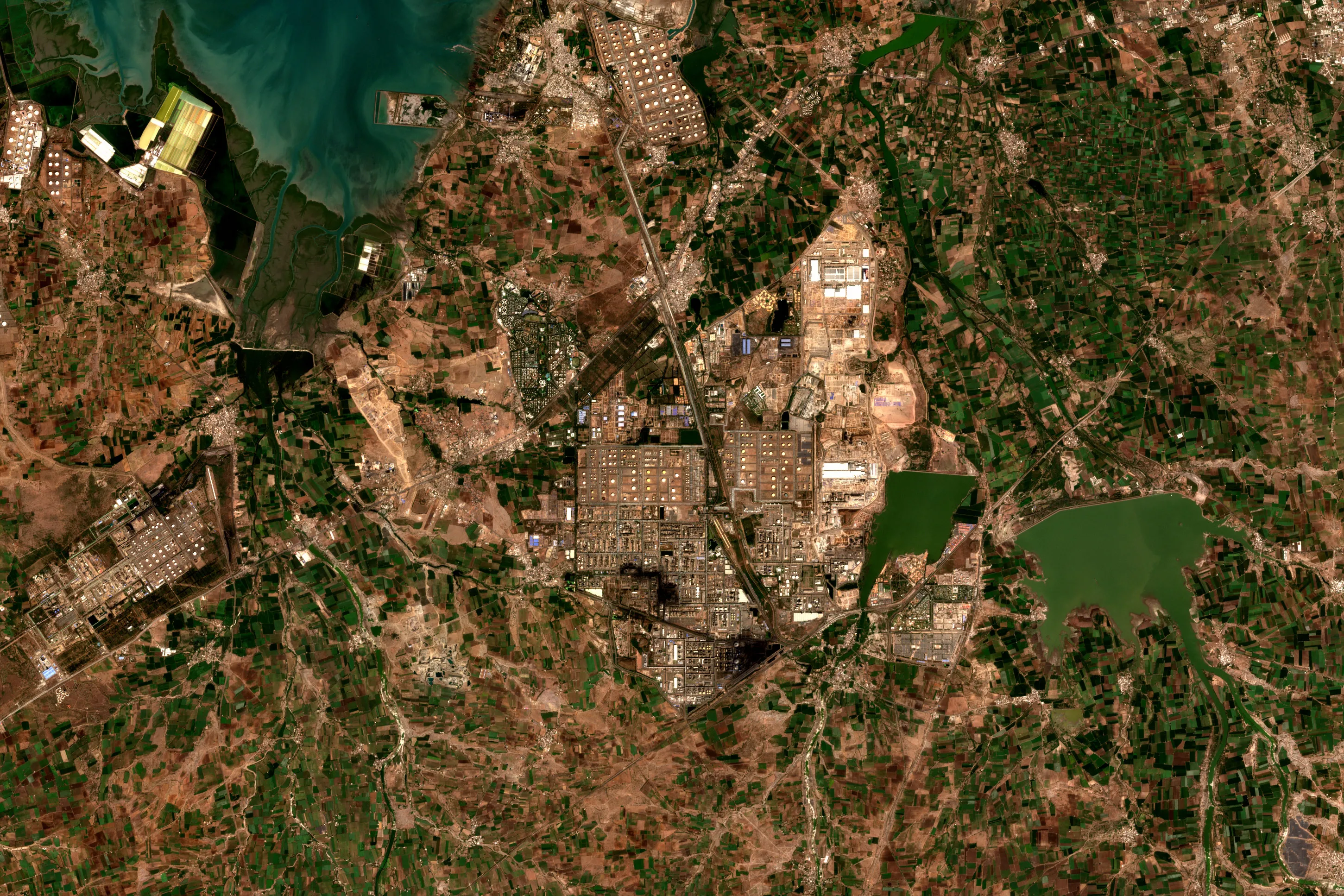 Satellite view of the Jamnagar refinery and petrochemical complex in Gujarat, India, with grids of storage tanks and process units among farmland, a green reservoir to the east and a turquoise bay to the north-west